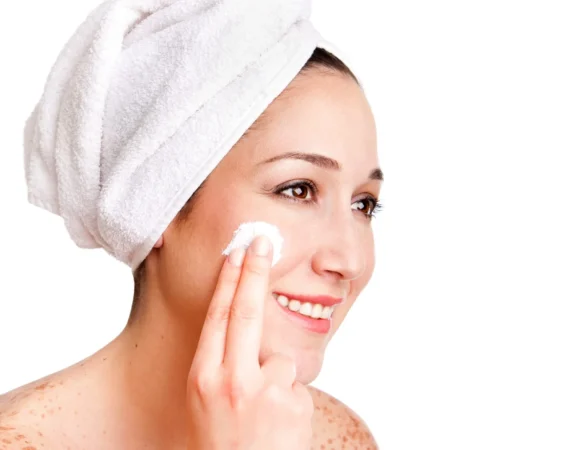 Smiling woman with a towel wrapped around her head applying cream to her cheek in a skincare routine.