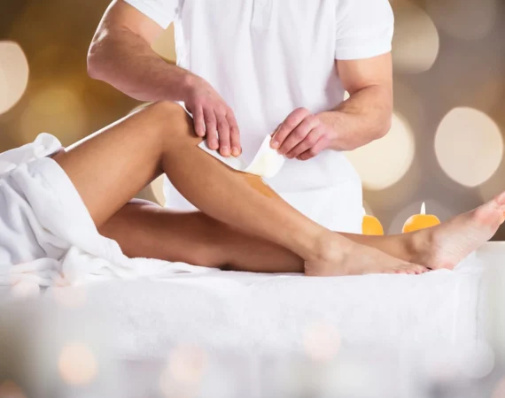 Spa therapist applying a wax strip to a client's leg during a waxing treatment on a white towel-covered table with candlelight in the background